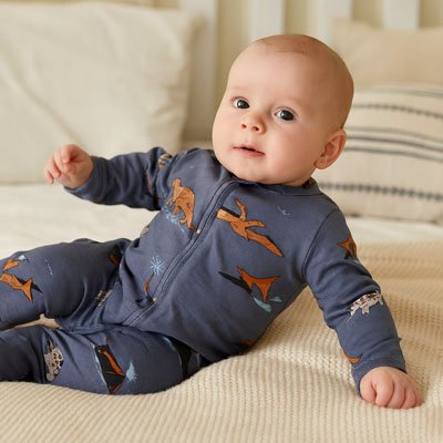 Bébé vêtu Pyjama bébé ultra-doux Coton Bio bleu foncé motifs dinosaures orange allongé sur couverture beige texturée à côté oreiller