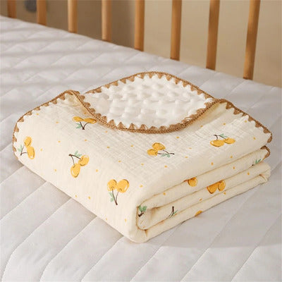 Couverture bébé Cocon Douceur en mousseline légère, motif cerises jaunes, pliée sur un matelas de berceau