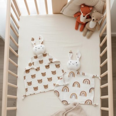 Deux Doudou Lapin Ultra-Doux Nuit Sereine Ourson et arc-en-ciel étalés sur lit bébé avec deux peluches en arrière plan