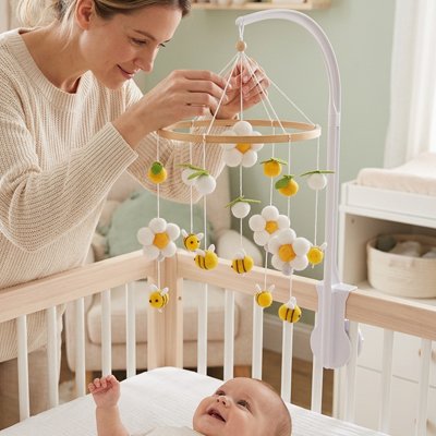 Vue profil gauche de bébé souriant dans son lit et regardant sa mère vêtu de beige fixer le mobile bébé apaisant musical suspendu au dessus de lui